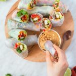 Wooden platter of 10 rice paper rolls with colorful vegetable fillings and a creamy orange dipping sauce.