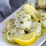 White cooked scallops with herbs and lemon wedges piled on a rectangular white plate with grey napkin in the background.