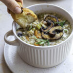 Mushroom rice soup with green garnishes in a white ramekin. A hand is dipping crusty bread into the soup from above.