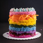 A tall round layered cake made of different colors of sushi rice and vegetables. There are layers of purple, blue, green, yellow, orange, pink and topped with piped blue and purple cream cheese.