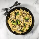 White table with black bowl of penne with broccoli florets and sliced mushrooms.