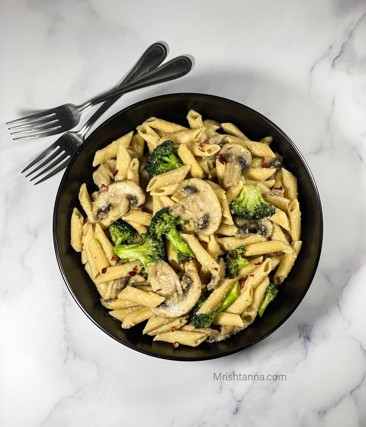 White table with black bowl of penne with broccoli florets and sliced mushrooms.