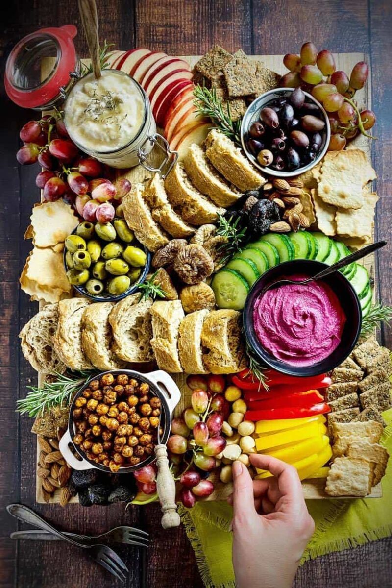 Very colorful board of vegan snacks including nuts, crackers, bread and dips including a bright purple dip in a black bowl surrounded by cucumber.