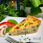 Slice of pale yellow quiche studded with mushrooms and spinach on a white plate with tomato and mushroom slices beside.