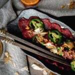 Deep purple red colored venison sashimi with sliced green jalapenos and bonito flakes on a dark grey dish.