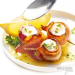 3 bacon wrapped scallops on a plate with melted butter, lemon wedge and chopped parsley.