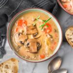 Bowl of pale brown orange creamy soup with mushrooms, tomatoes and green herbs in it.