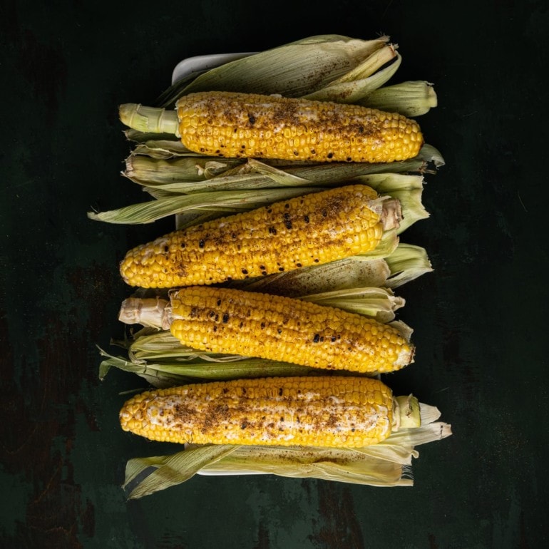 Black background with 4 ears of roasted corn on the cob set on their pale green husks.