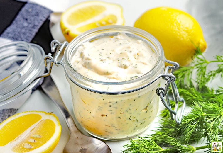 Glass canning jar of green flecked creamy tartar sauce. There are lemons and dill surrounding the jar. 