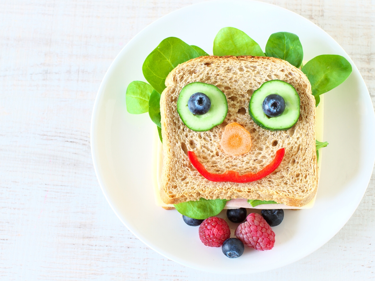 Whole wheat sandwich decorated to look like a face with cucumber and blueberry eyes, red pepper slice mouth and a carrot nose.