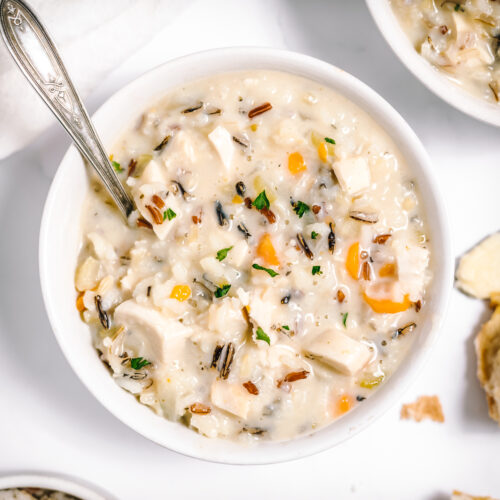 Creamy soup with flecks of brown wild rice, white chicken and orange carrots in white bowls on a white table. Silver colored spoon is in the bowl on the left side.
