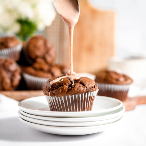 A spoon dripping pale brown cinnamon glaze onto a brown gingerbread muffin on a stack of small white plates.