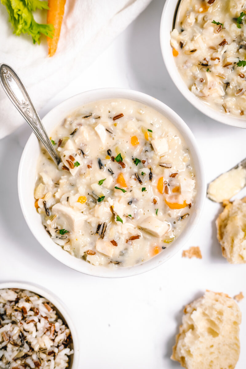 Creamy soup with flecks of brown wild rice, white chicken and orange carrots in white bowls on a white table.
