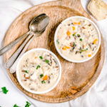 2 bowls of creamy white soup showing chunks of chicken, carrot and wild rice on a wooden round serving board with spoons beside. Square photo.