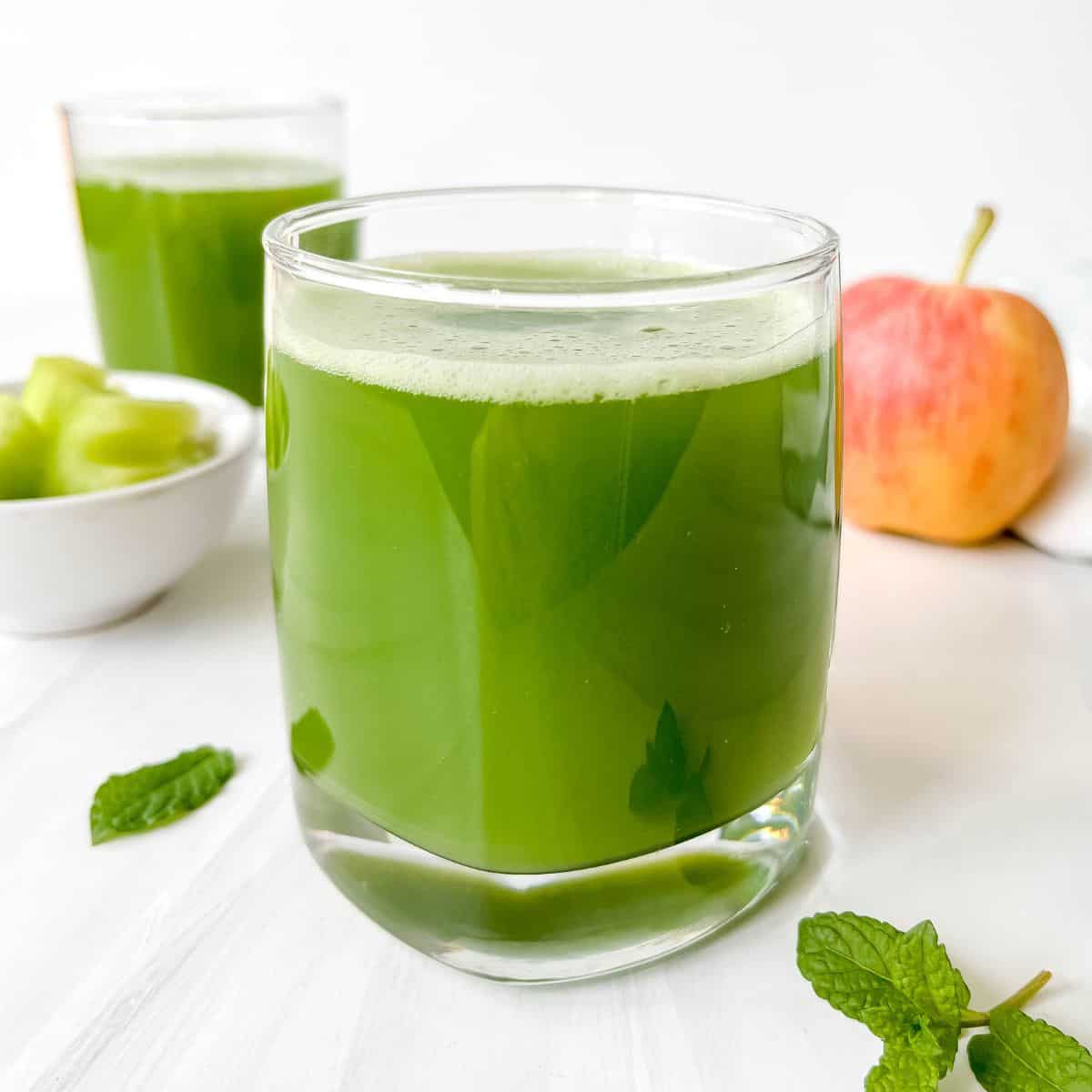 2 glasses of green juice with an apple, mint and cucumber slices around them.