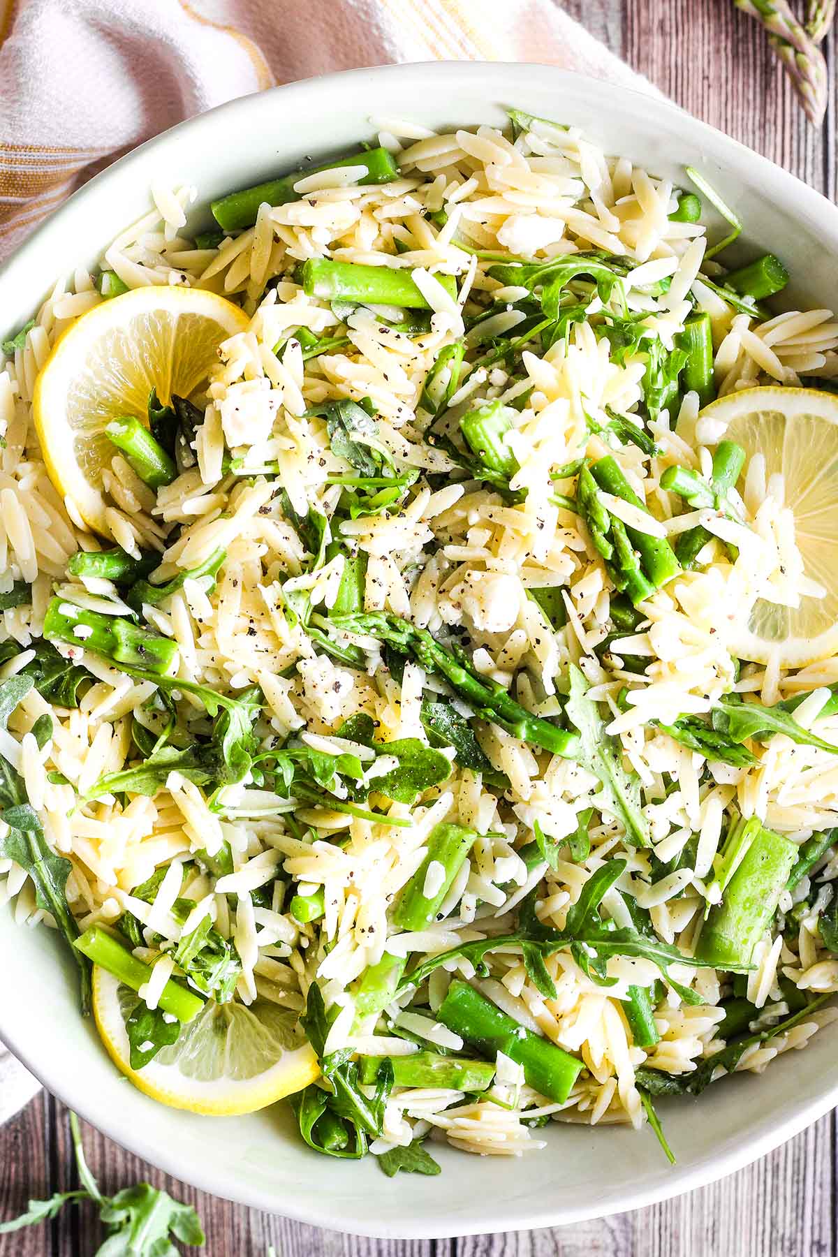 White bowl of small orzo pasta with chopped asparagus, arugula and lemon slices in it.