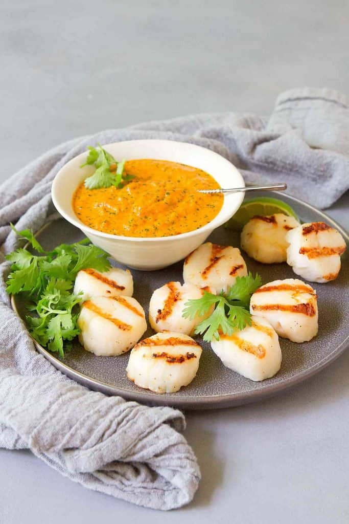 White scallops with brown grill marks on a grey plate with basil leaves around and a dish or bright orange dipping sauce.