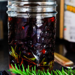 Mason jar with dark purple blackberries and vinegar and herb sprigs in it. It's on a black counter top with a sprig of rosemary in front.
