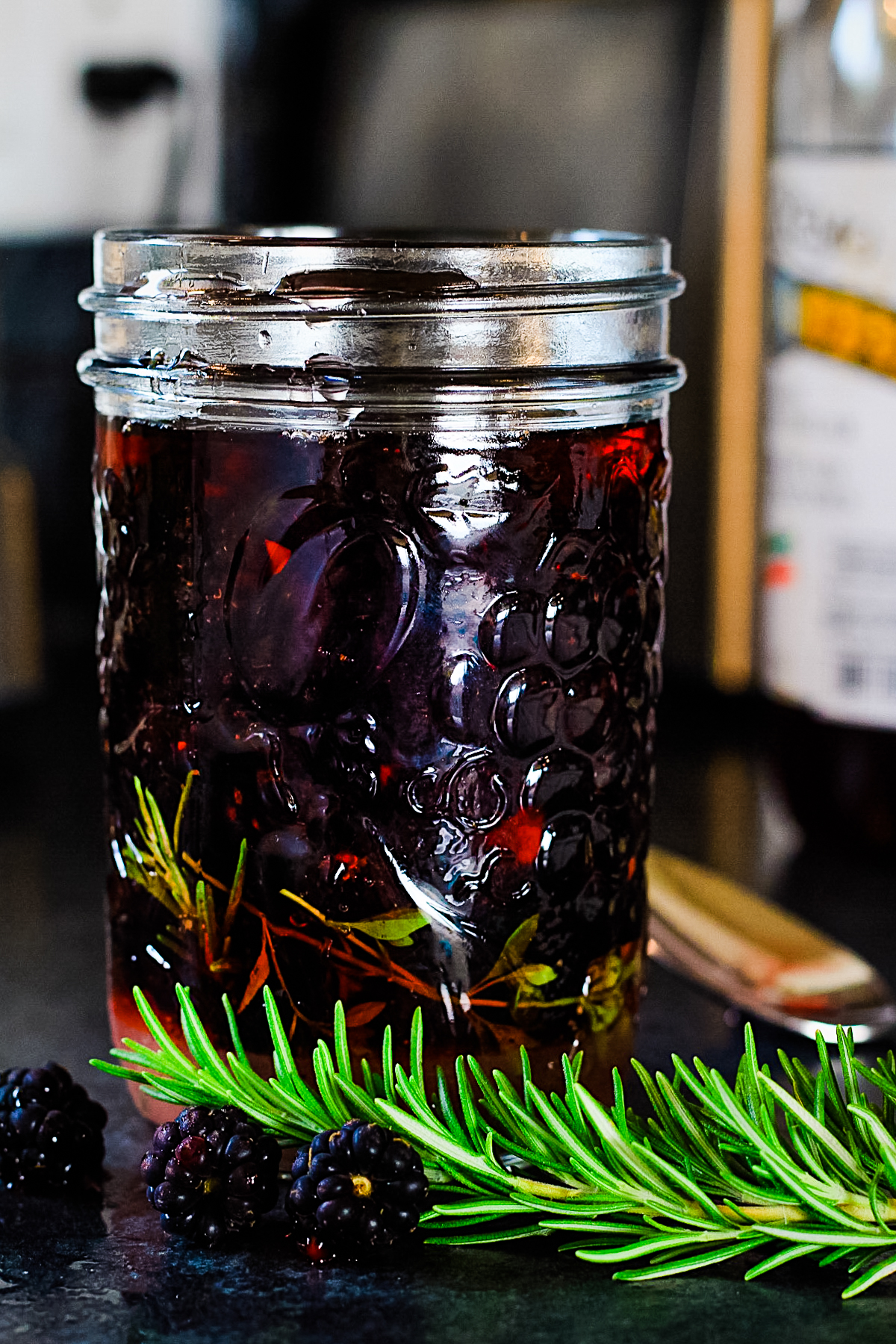 Mason jar with dark purple blackberries and vinegar and herb sprigs in it. It's on a black counter top with a sprig of rosemary in front.