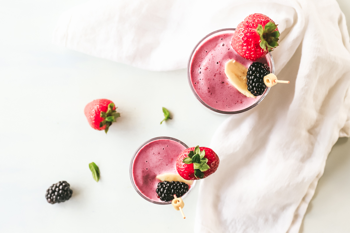 Two glasses full of bright purple fruit smoothie mixture garnished with skewers of banana, strawberry and blackberry. There are a few mint leaves and berries scattered on the table around the glasses.
