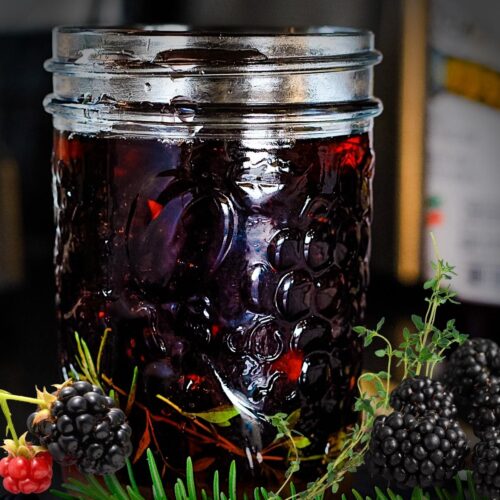 Mason jar of dark purple pickled berries surrounded by fresh blackberries and herbs.