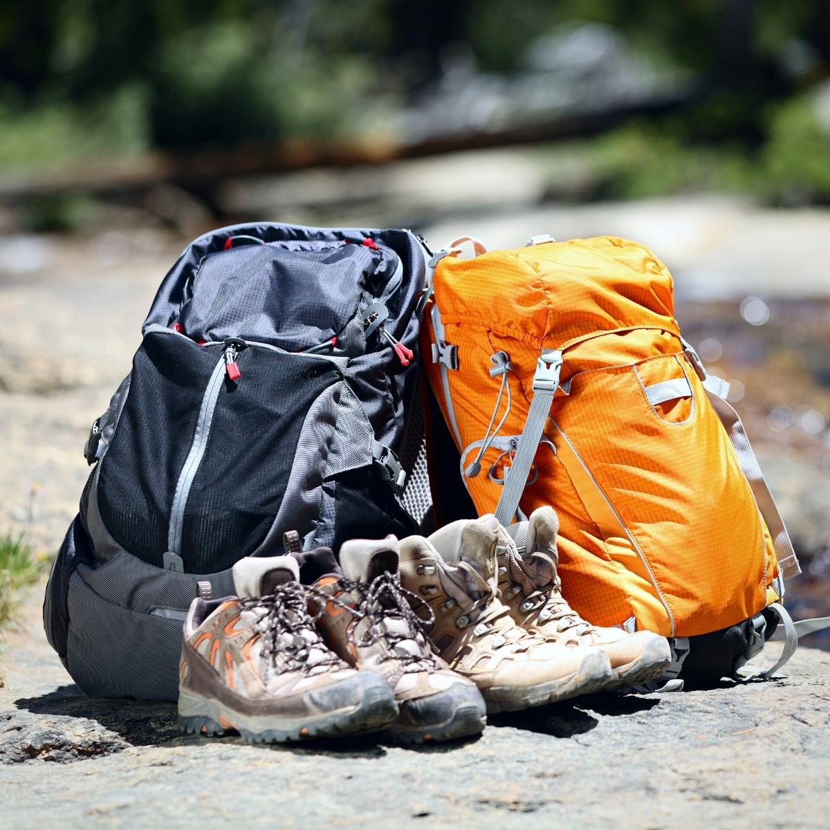 An orange backpack and black backpack side by side with 2 pairs of hiking boots in front.