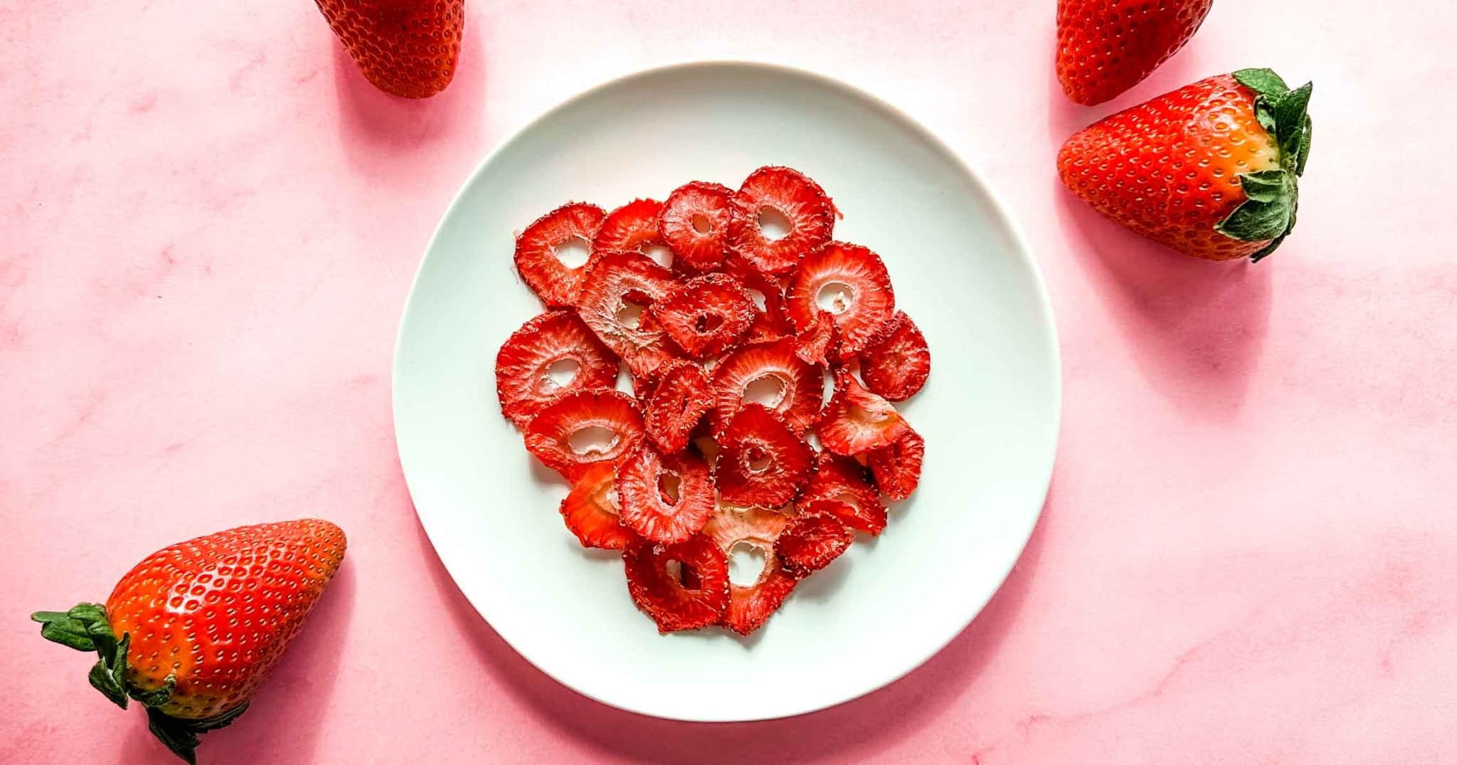 Dried sliced strawberries on a white plate with fresh strawberries around on a pink table.