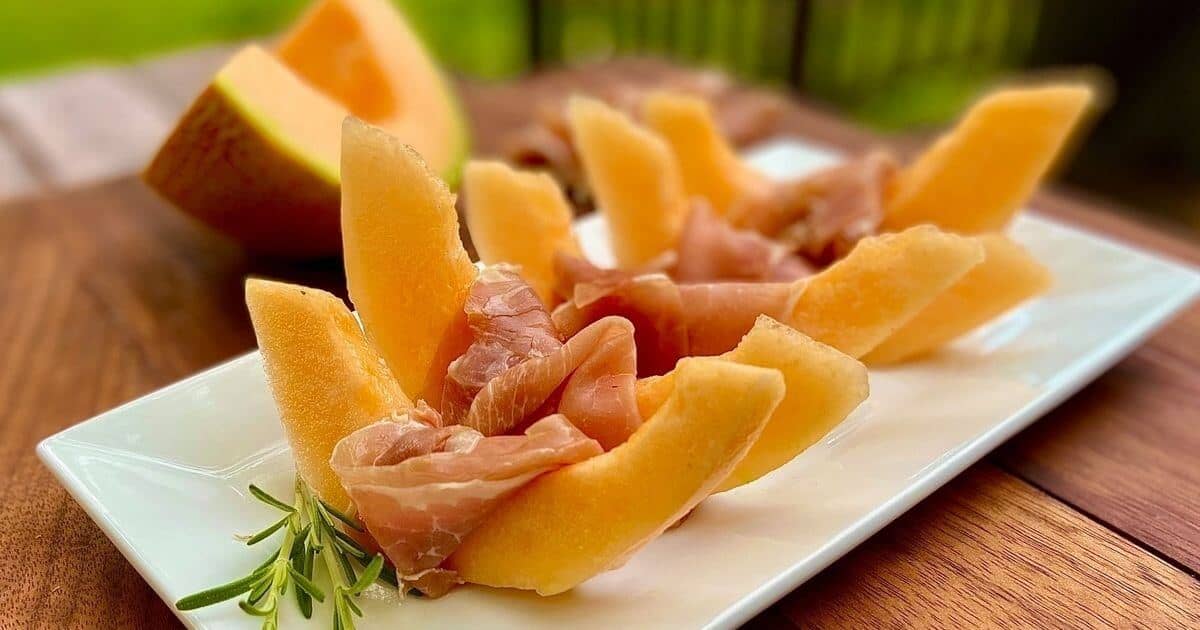Wedges of cantaloupe melon with slices of thin prosciutto draped over the middle on a long white platter. 