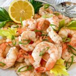 A pile of shrimp with oil, lemon and herbs drizzled over and a lemon half beside.