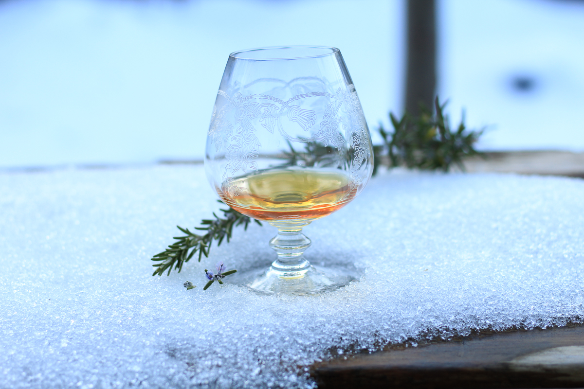 An etched goblet with scotch in the snow with a sprig of rosemary beside.