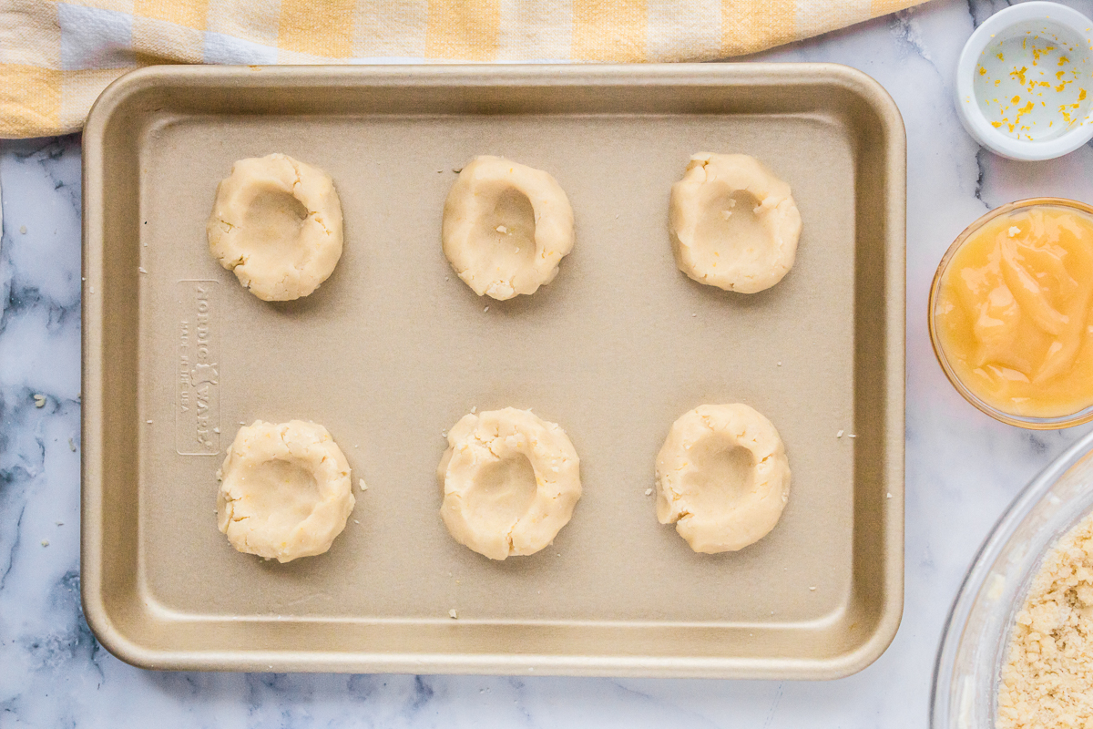 Dough has been formed into small round disks with indents in the middle on a beige baking sheet.