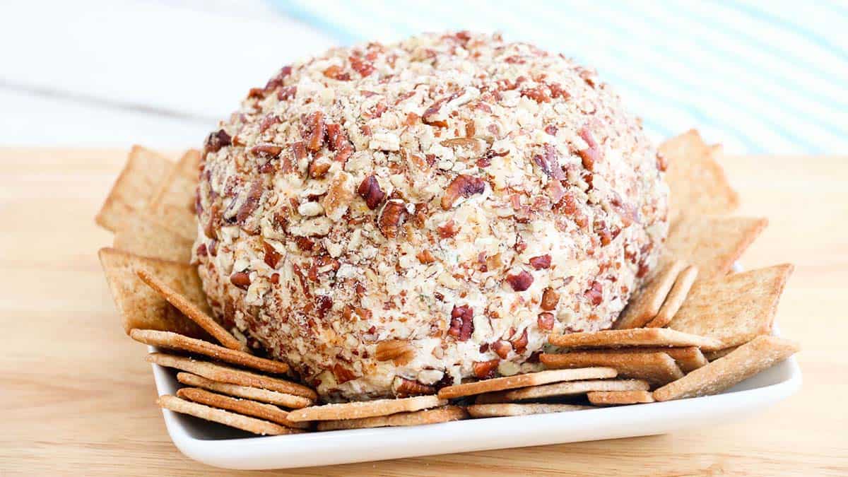 A ball of cheese mixture coated in light brown crushed nuts and surrounded by square wheat crackers.