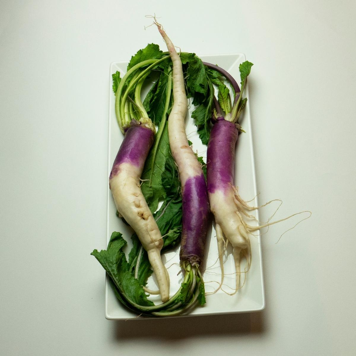 Long purple and white turnips with their greens on a white board.