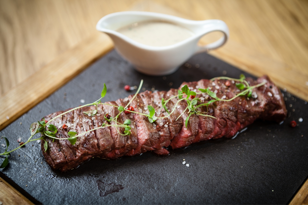 A grilled and sliced flank steak garnished with herbs on a black square board with a white sauceboat of creamy sauce beside.