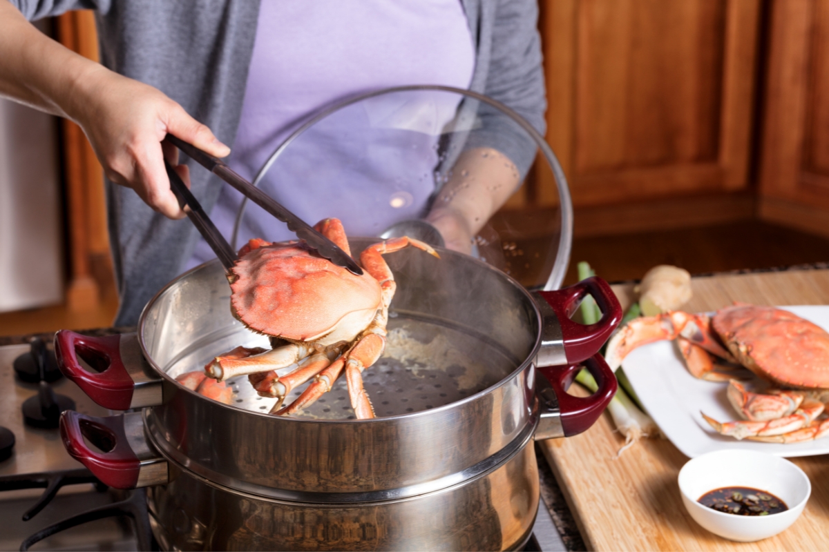 A hand with tongs lifting a whole bright pink crab out of a silver pot of boiling water.