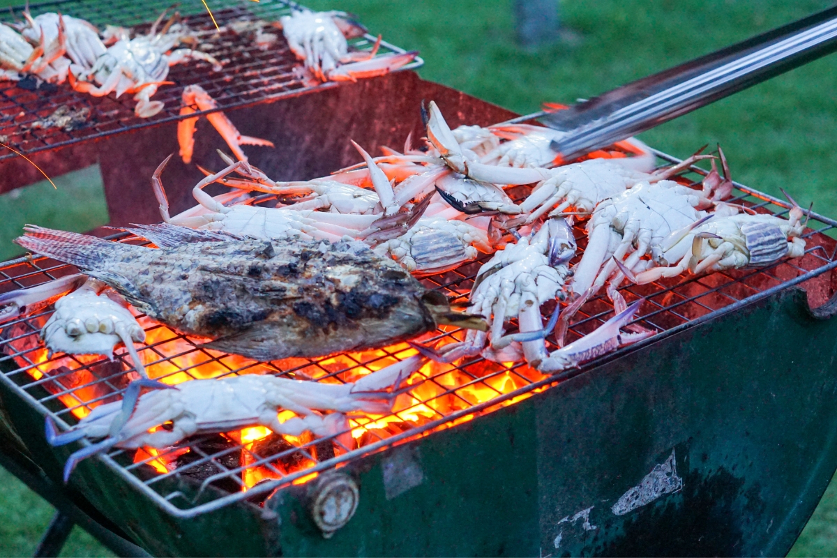 A grill of glowing coals with crabs and fish on top and tongs near to flip the seafood.