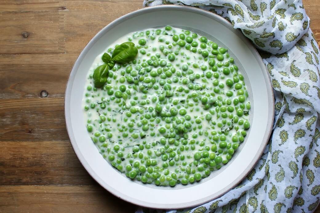 Creamed green peas in a pottery bowl on a medium brown wooden counter with a patterned blue tea towel beside.