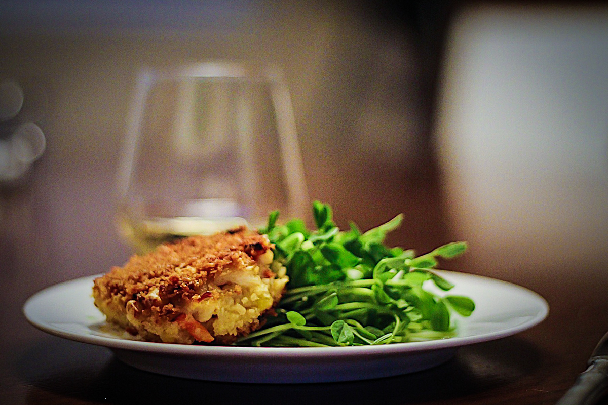 White plate with a crispy breaded crab cake sitting on a green salad with a stemless glass of white wine in the background.
