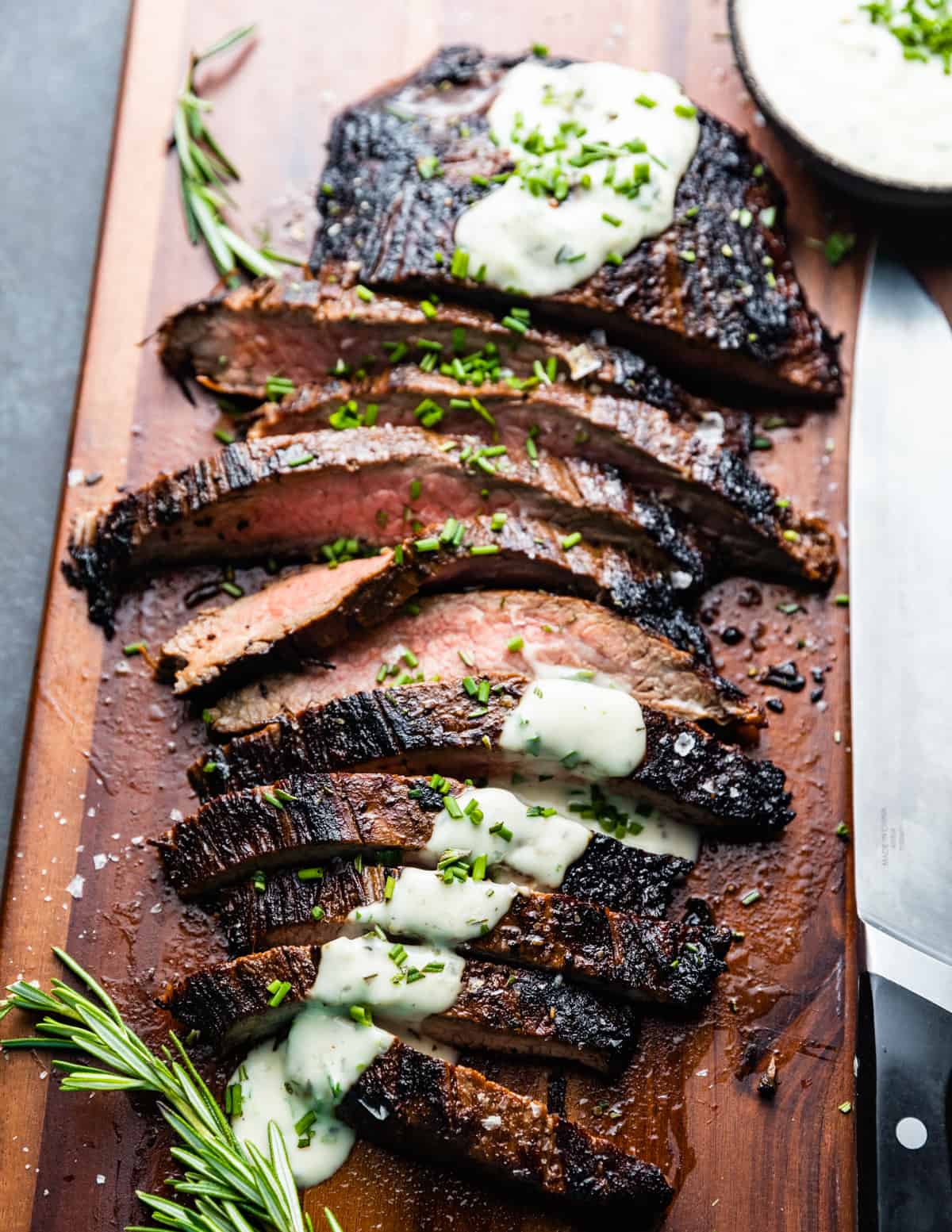 Well seared and sliced steak on a wooden cutting board. There is white sauce napped over the top in a stripe and green herbs garnishing.