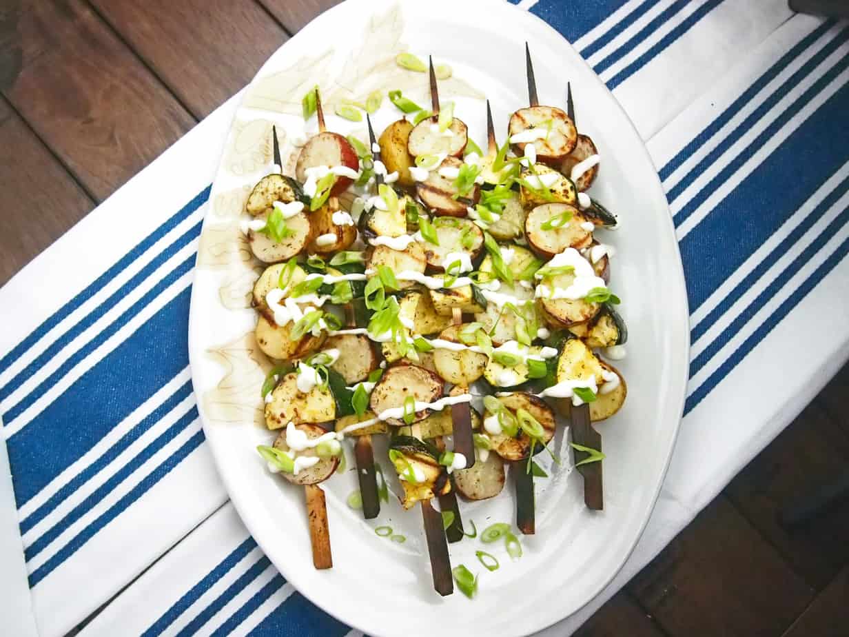 BBQ'd potato salad skewers are white and green with charred skewers. All on a white oval platter drizzled with white sauce.