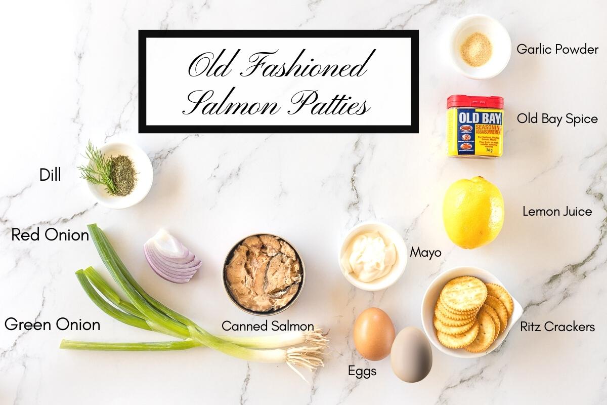 Labelled ingredients for old fashioned salmon patties recipe. Shown is dry dill weed, green onions, red onion, an open tin of canned salmon, mayonnaise, eggs, round crackers, a lemon, old bay seasoning and garlic powder.