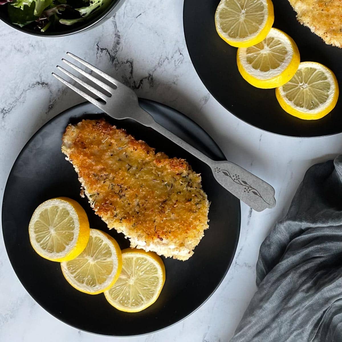 Crispy chicken cutlet with 3 slices of lemon on a black plate.