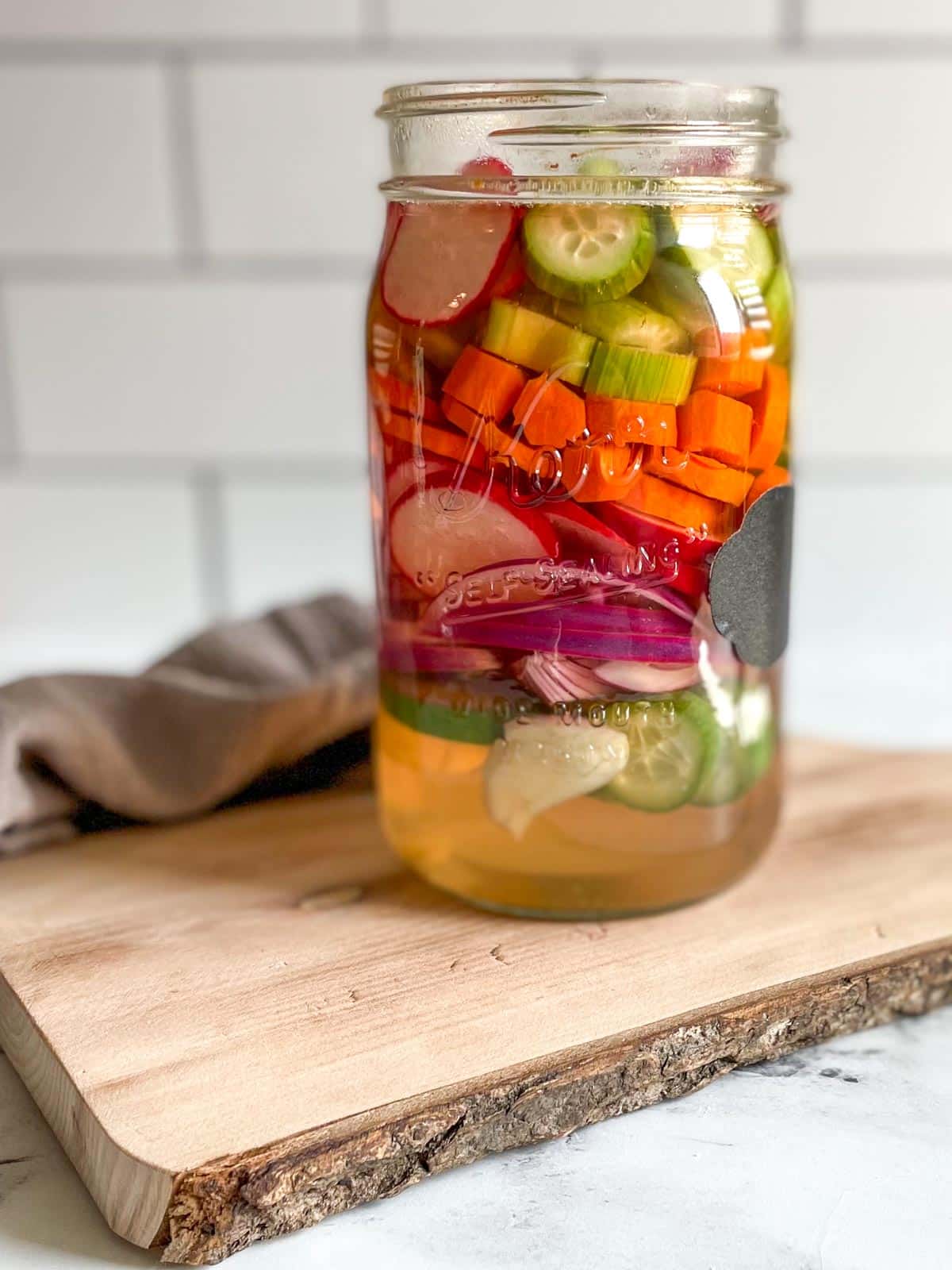 A tall mason jar layered with colorful vegetables floating in a clear pickling liquid.