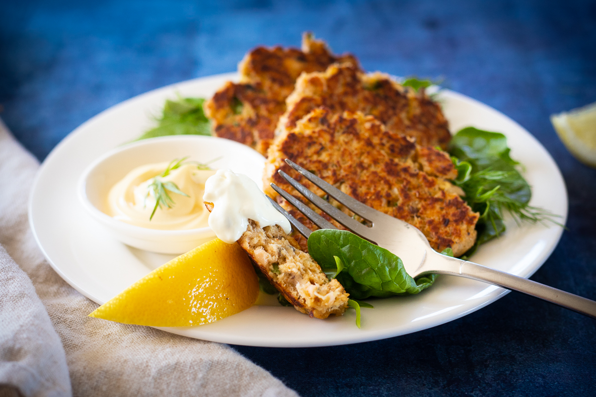 Pale pink salmon cakes recipe on green lettuce leaves on a white plate with creamy white dip and a yellow lemon wedge. 