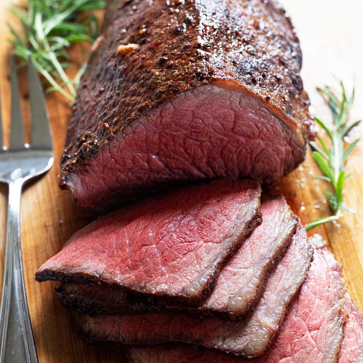 Rare roasted beef with a brown crust being sliced on a wooden cutting board.