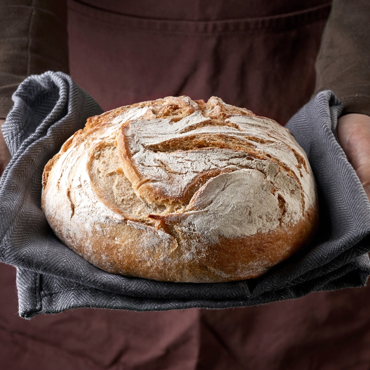 A brown and round loaf of bread being held by hands in a grey tea towel. There is a light dusting of white flour over the bread.