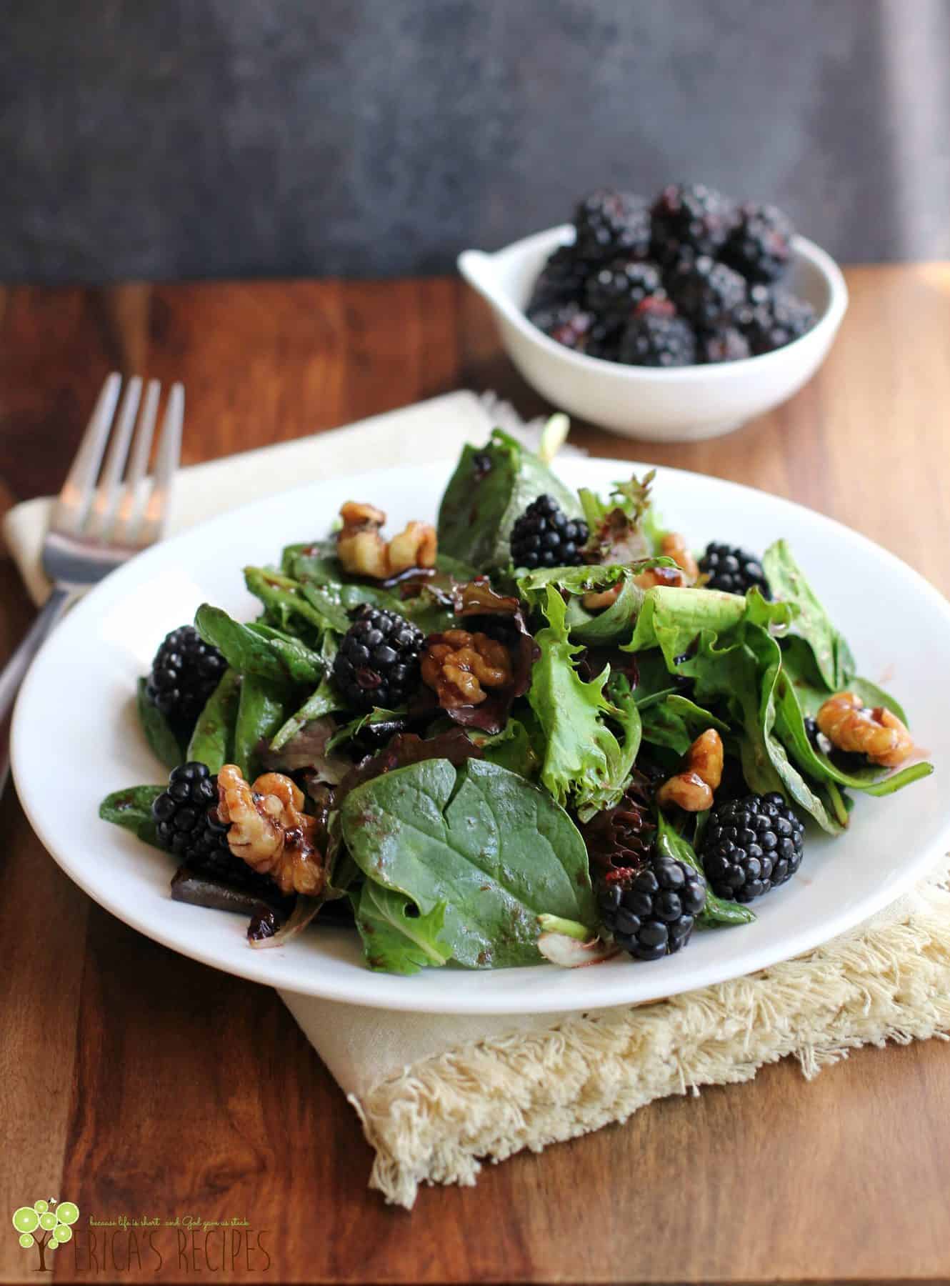 A dark green salad with nuts and blackberries scattered through and a small white dish of fresh blackberries in the background.