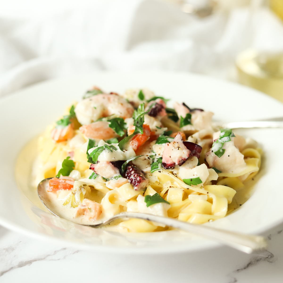 White wide rimmed bowl filled with red, white and purple assorted seafood chunks over fettuccini pasta.