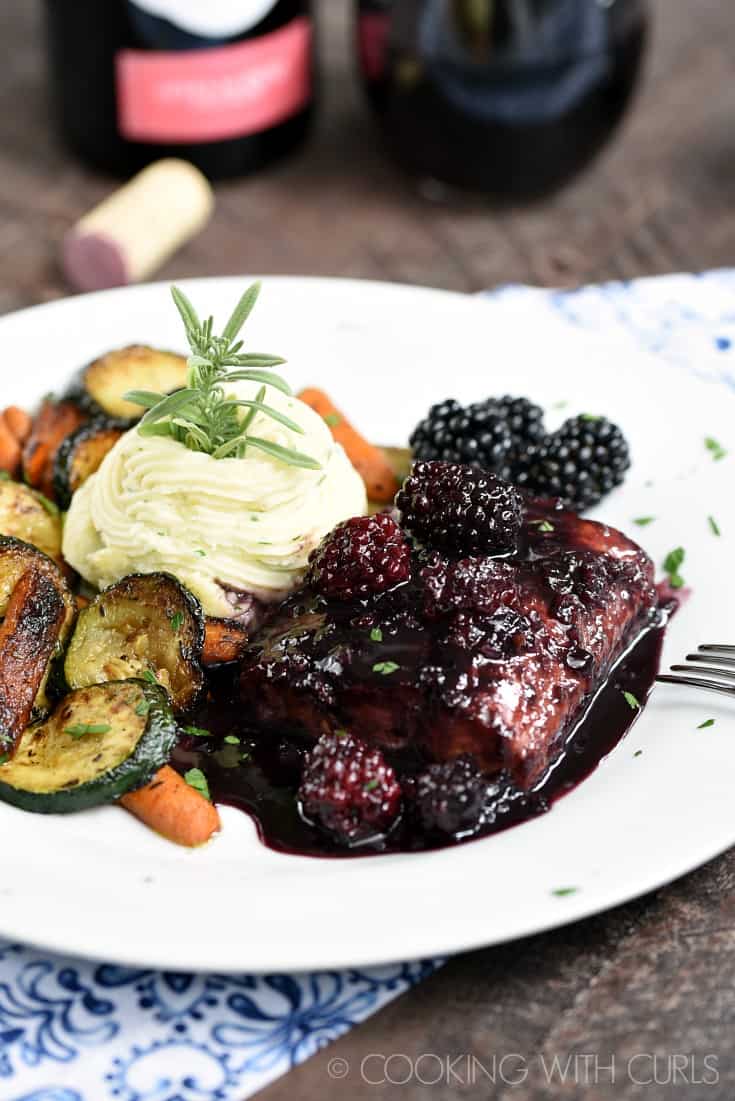 Dark purple blackberries and sauce over a serving of salmon on a plate with creamy white mashed potatoes and grilled carrots and zucchini.