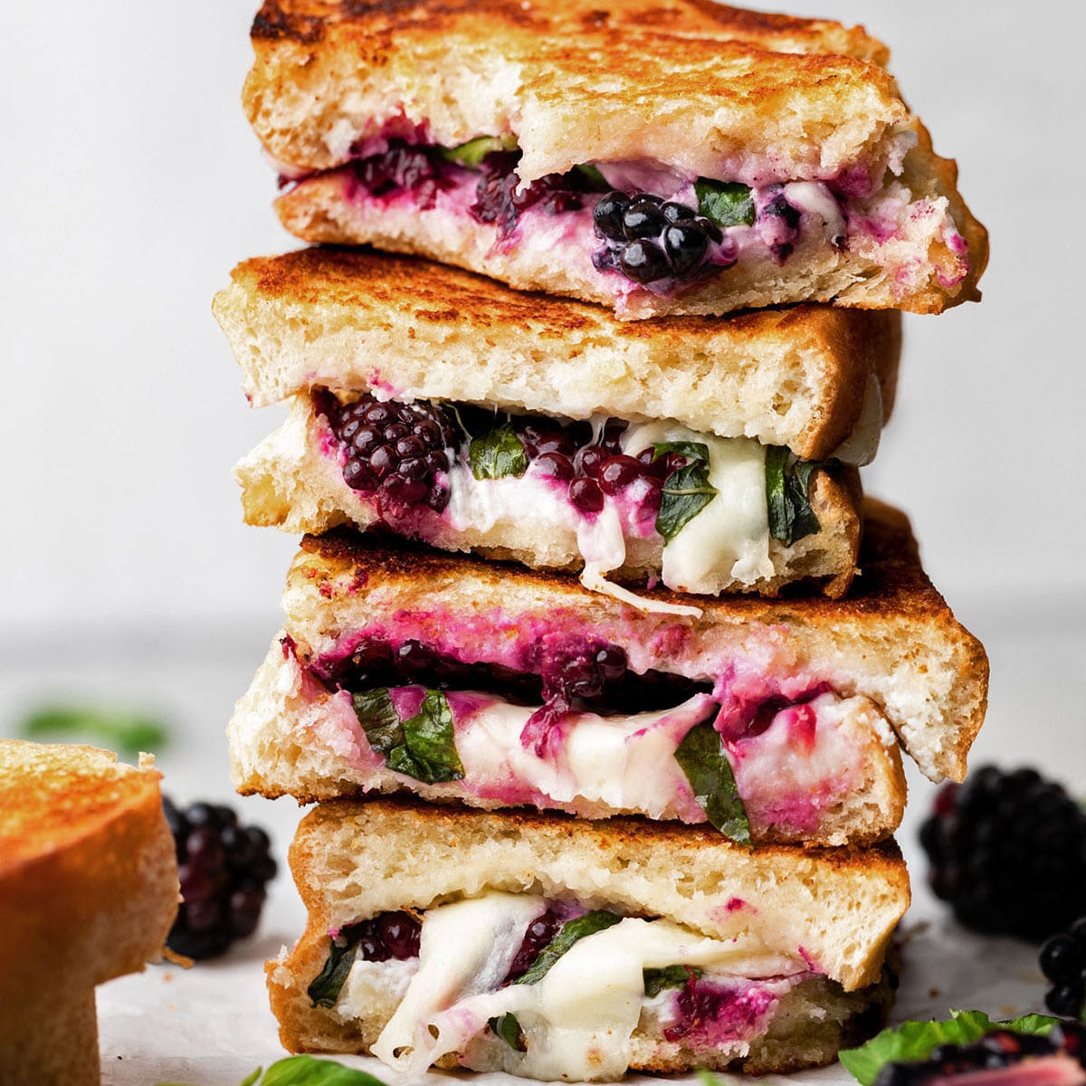 A stack of 4 sourdough grilled cheese and blackberry sandwiches on a plate. You can see the berry juice and melted cheese dripping out of them.